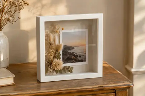 Cadre photo vitrine carré blanc posé sur une console, présentant une photographie et des fleurs séchées
