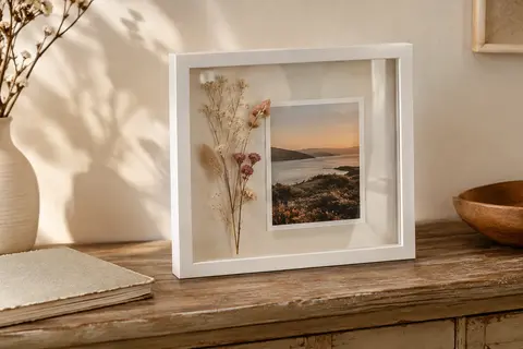 Cadre photo 3D blanc posé sur une console, avec fleurs séchées et photographie visibles derrière la plaque transparente.